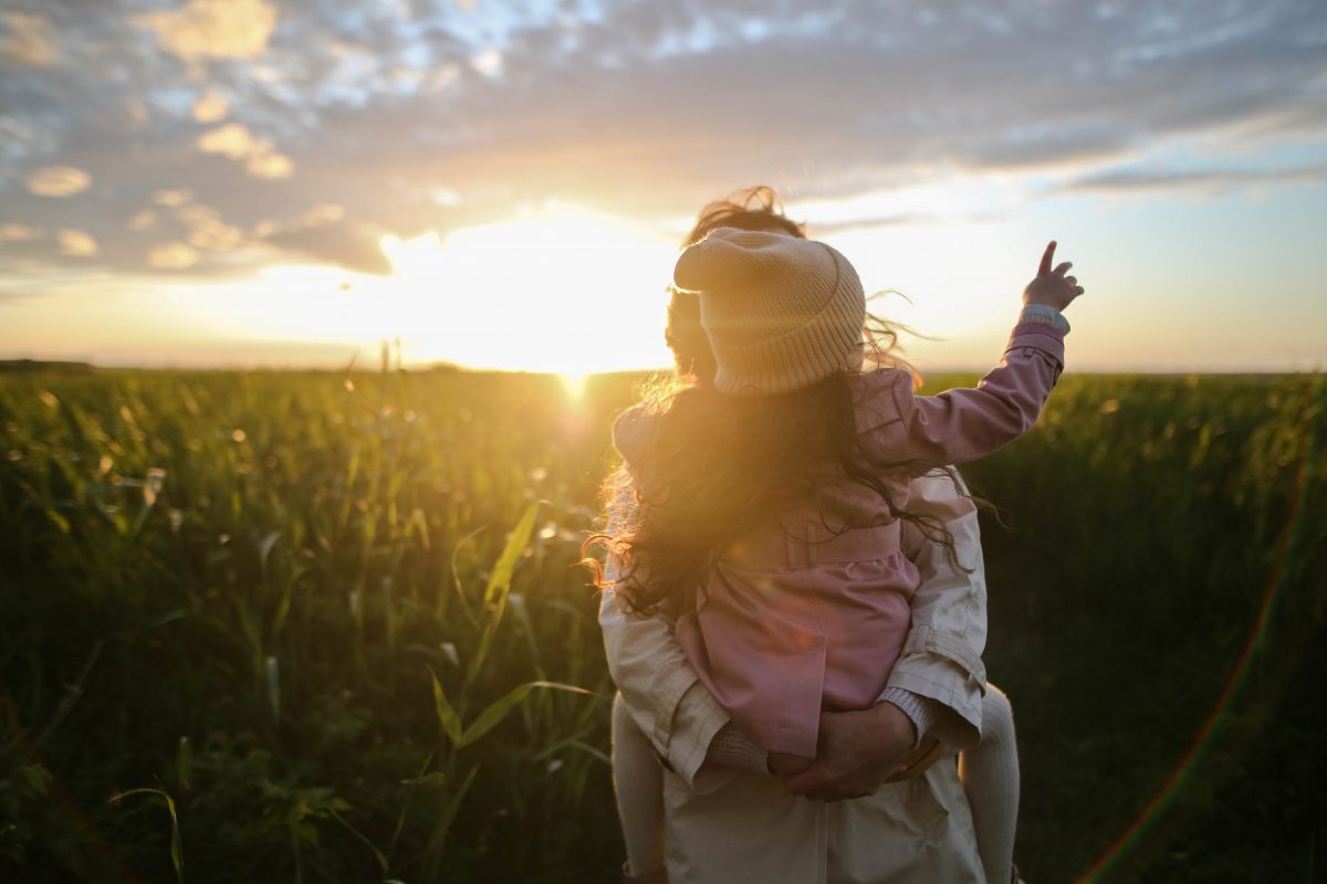 Femme brune tenant une petite fille dans les bras dans un champ. Le soleil se couche. La petite fille montre le soleil.